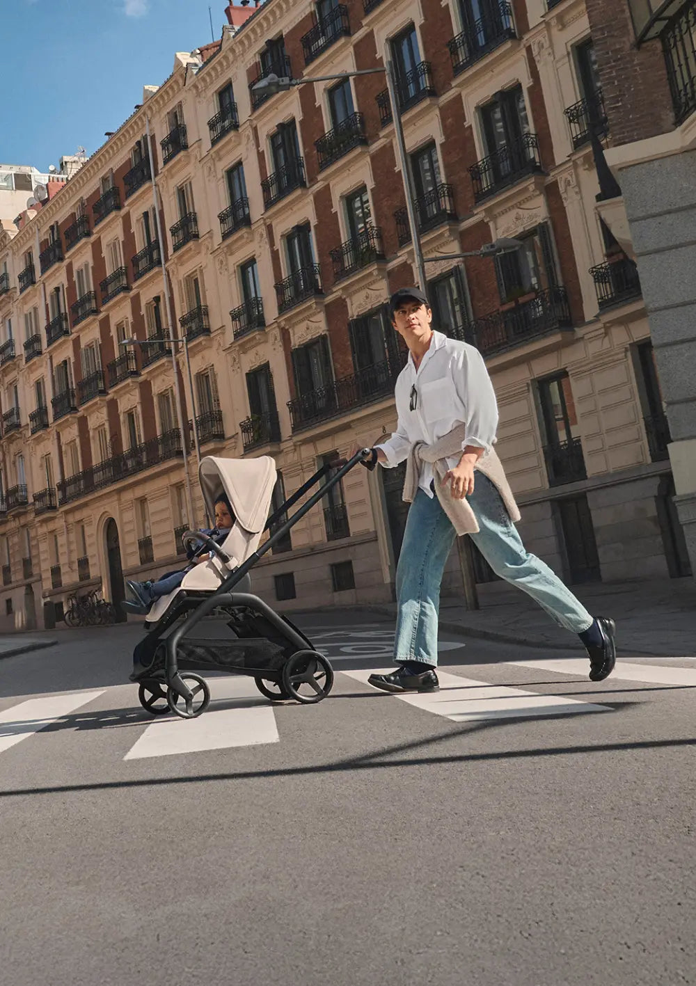 Ein Mann schiebt den kompakten Bugaboo Kinderwagen Dragonfly Plus mit Sportsitz über einen Zebrastreifen in einer historischen Stadtstraße; idealer Begleiter für urbane Wege.