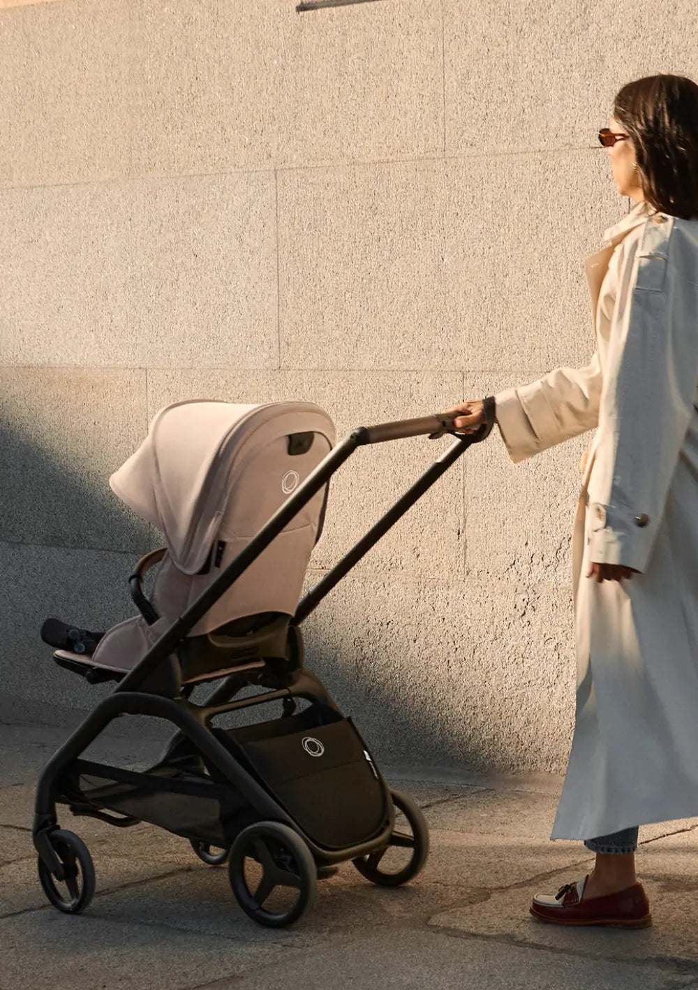 Eine Frau im eleganten Trenchcoat schiebt den stilvollen Bugaboo Kinderwagen Dragonfly Plus an einer sonnenbeschienenen Steinwand entlang; Blick auf die Rückseite des Sitzes und das schwarze Fahrgestell.