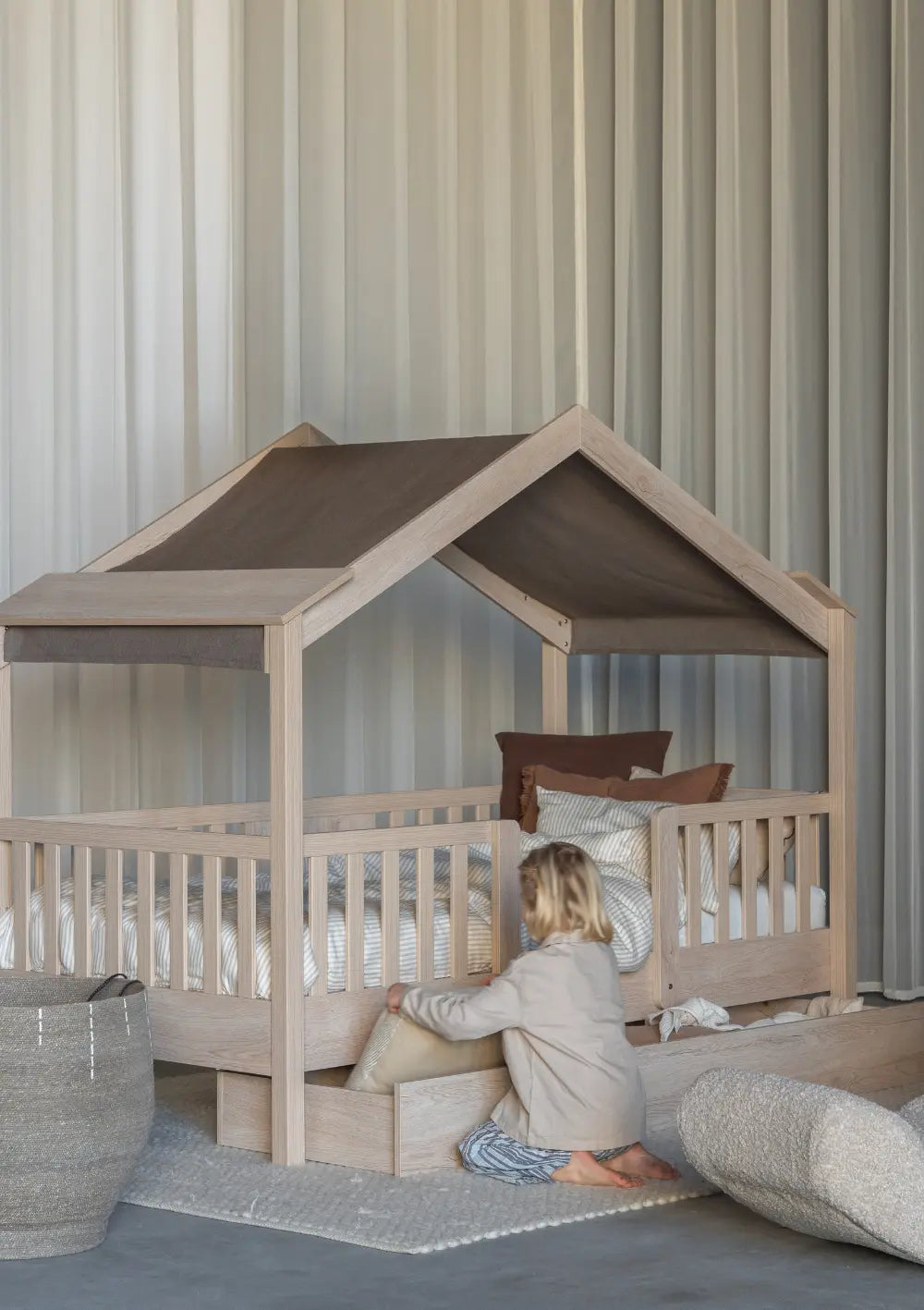 Ein Kind öffnet die ausziehbare Bettschublade (separat erhältlich) unter dem Quax Hausbett Chalet Natural Ash (200 x 90 cm), welches hier mit einem gemütlichen, dunkelbraunen Stoffdach (Farbe Chocolate) als Betthimmel ausgestattet ist.