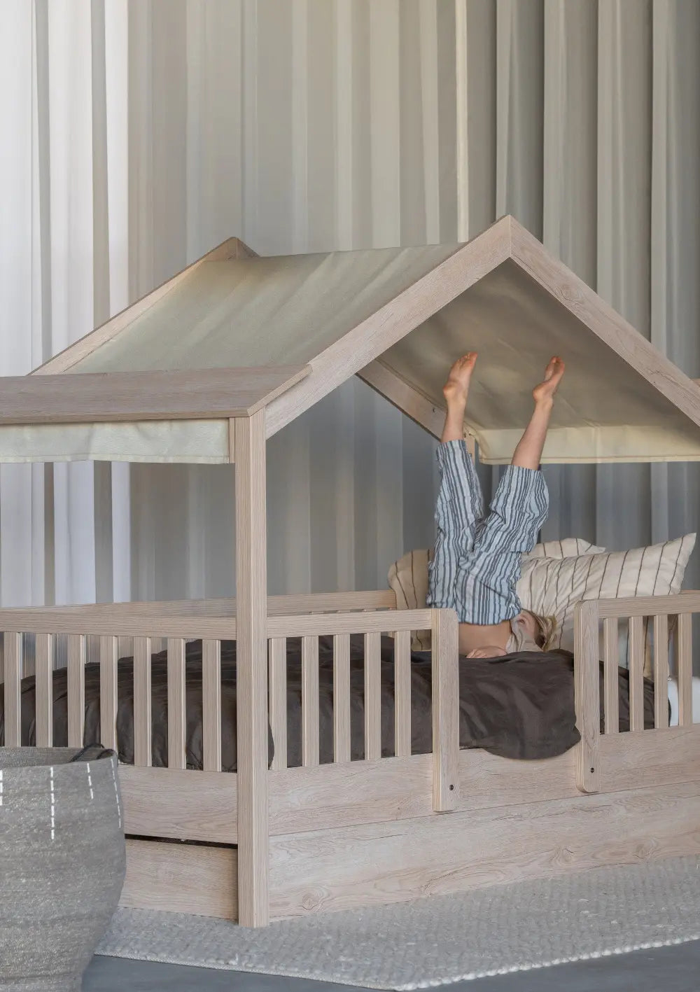 Ein Kind macht einen spielerischen Kopfstand im Quax Hausbett Chalet Natural Ash (200 x 90 cm), dessen Dachgestell mit einem hellen, cremefarbenen Textilhimmel (Farbe Butter) bespannt ist.