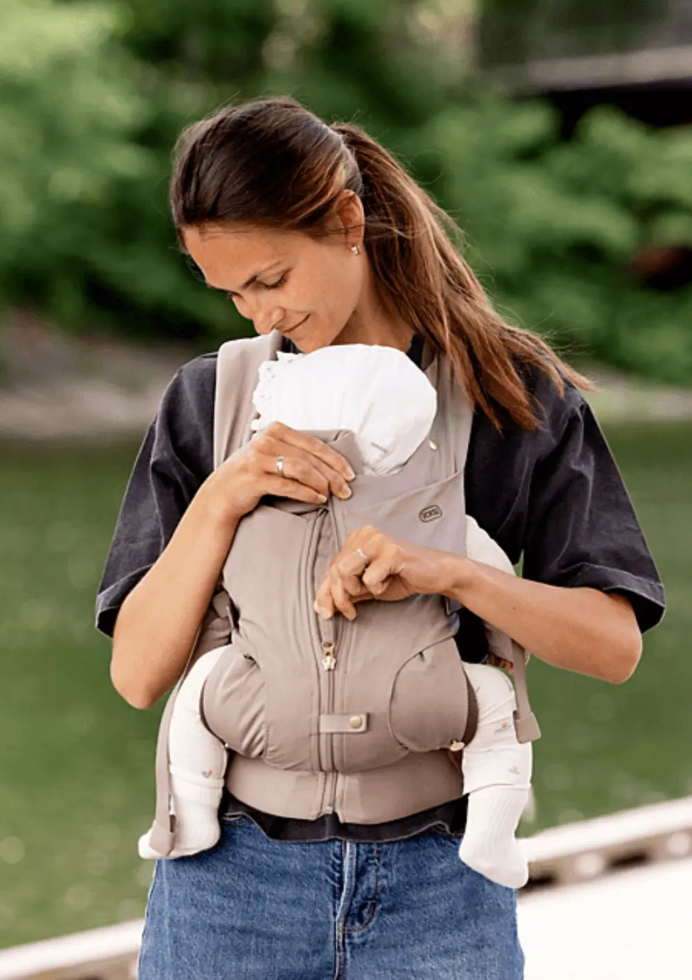 Eine Frau verstellt am Steg der Voksi Babytrage Huddle Clay den praktischen Reißverschluss zur idealen Anpassung an die Ergonomie des Kindes; im Hintergrund ein Gewässer.