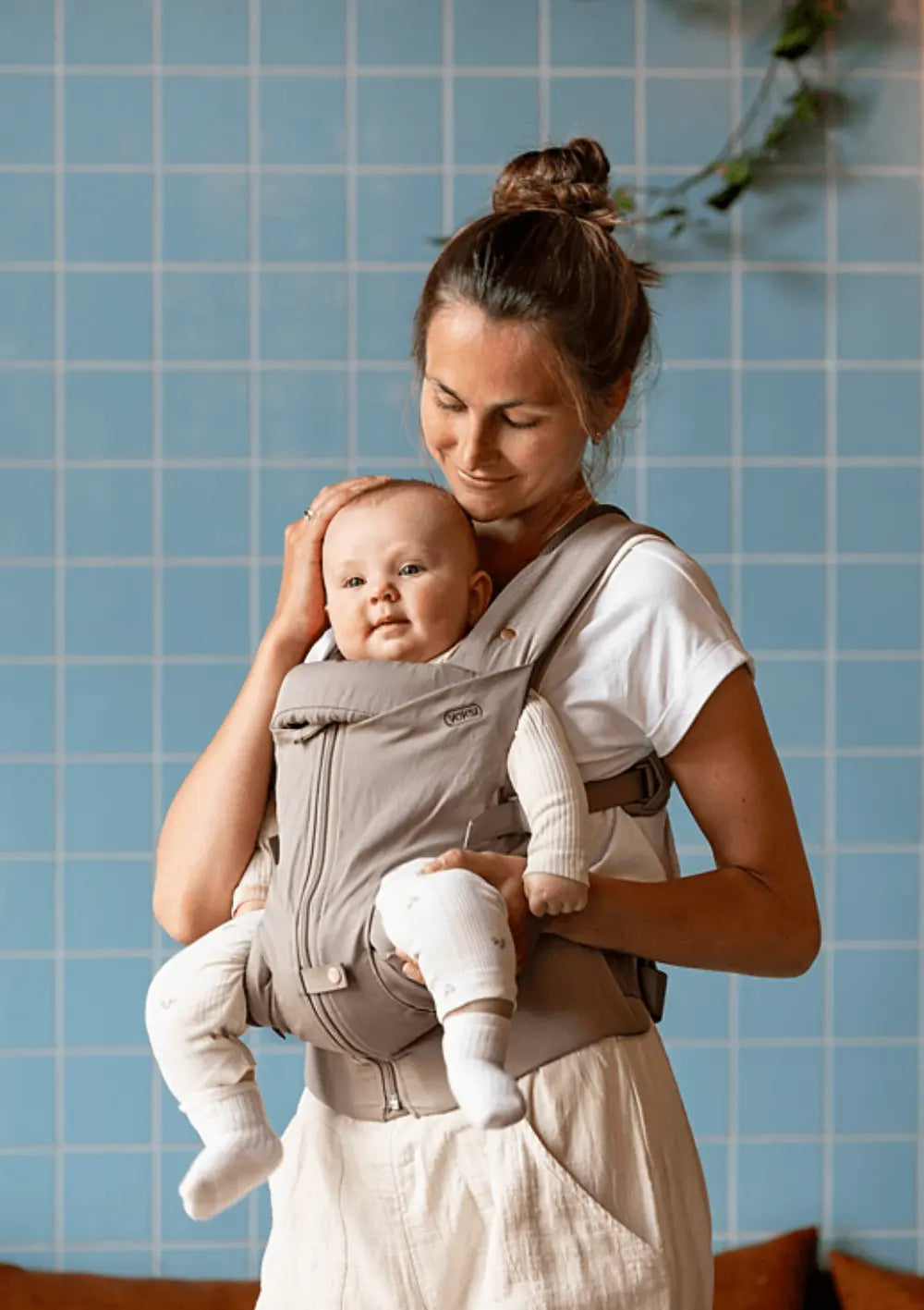 Eine Mutter blickt lächelnd auf ihr Baby, das sie in der beigeschlamm-farbenen Voksi Babytrage Huddle Clay vor dem Bauch trägt; hellblaue Fliesenwand im Hintergrund.