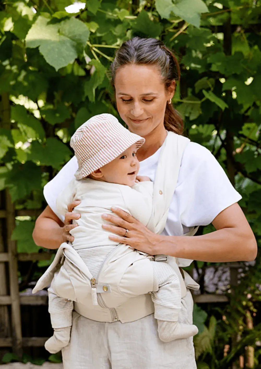 Eine Mutter blickt lächelnd auf ihr Baby mit Sonnenhut, das sicher in der cremefarbenen Voksi Babytrage Huddle Cream vor ihrer Brust sitzt; im Hintergrund grüne Blätter.