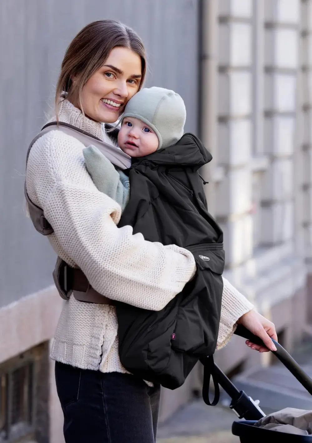 Eine lächelnde Frau hat ihr Kleinkind in einer ergonomischen Komforttrage vor dem Bauch angelegt; das wärmende Voksi Cover für Babytragen Black schützt den Körper des Babys vor Kälte.