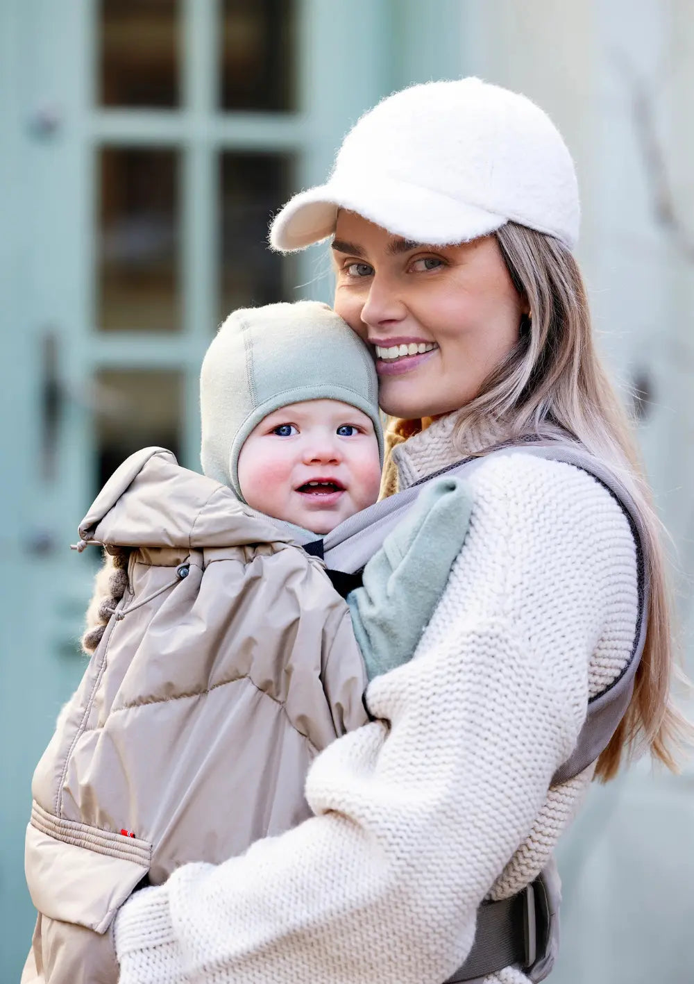Eine lächelnde Frau trägt ihr Kleinkind in einer Babytrage vor dem Bauch; das sandfarbene, wärmende Voksi Cover für Babytragen Dark Sand schützt das Kind beim Spaziergang vor Wind und Kälte.