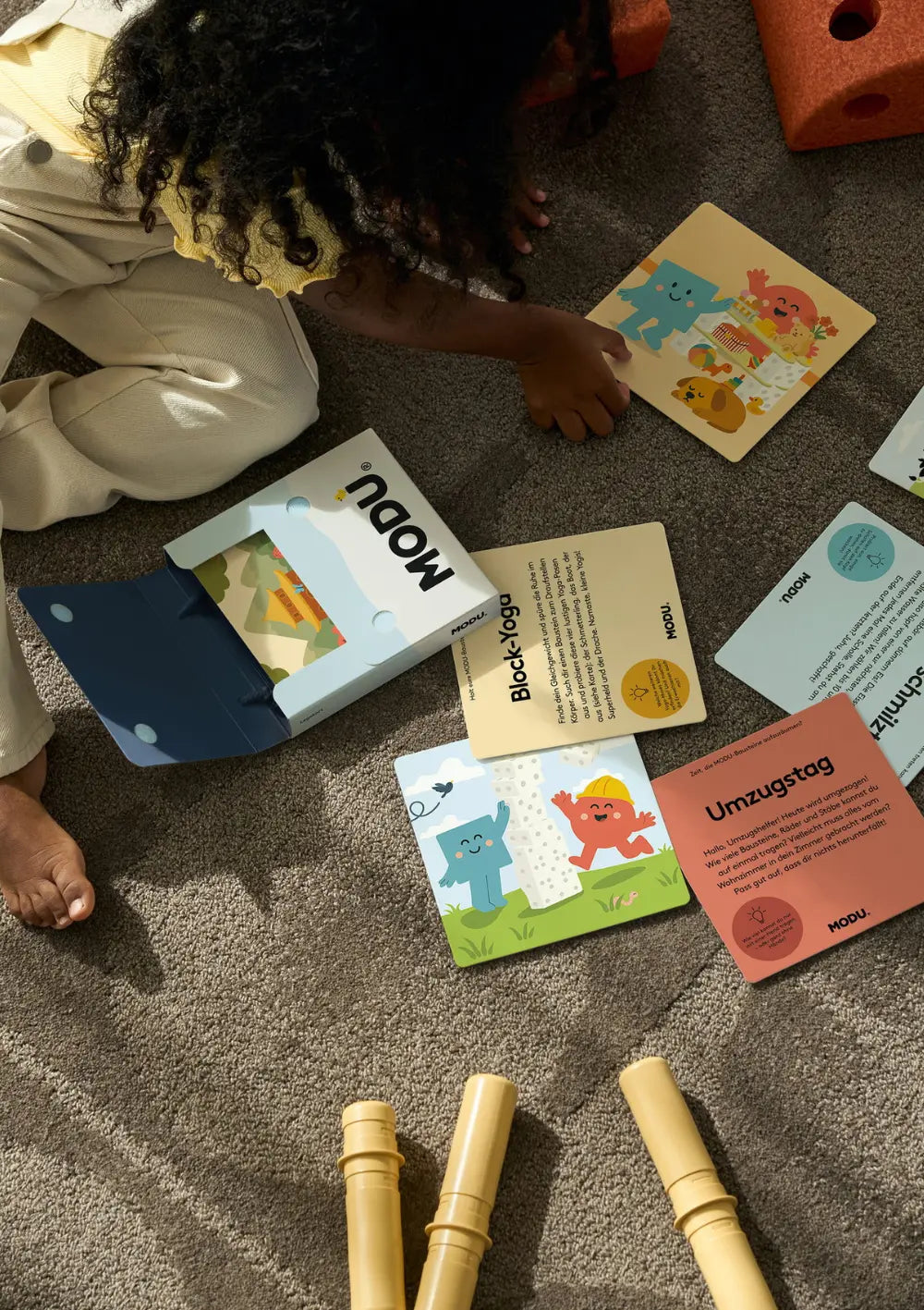 Auf einem Teppich liegen die geöffnete Verpackung und verschiedene, farbenfrohe Karten aus dem MODU Spielkarten-Set mit deutschen Spieletiteln wie „Block-Yoga“ und „Umzugstag“.