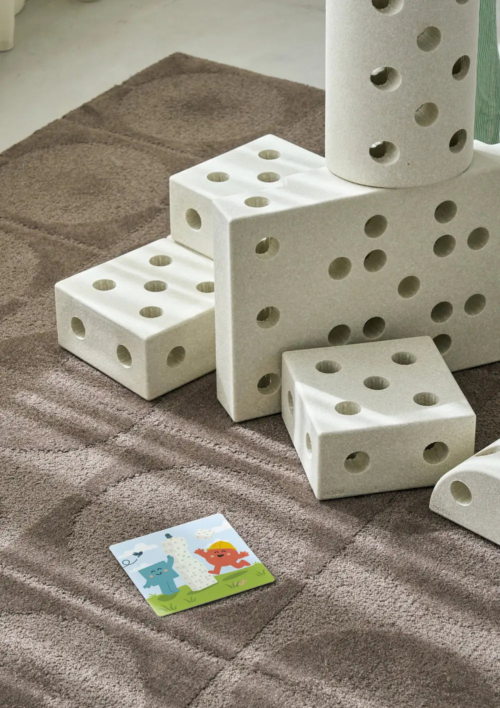 Eine kleine, illustrierte Aktivitätskarte des MODU Spielkarten-Sets liegt auf dem Teppich vor einer selbstgebauten, treppenartigen Skulptur aus hellen MODU Schaumstoff-Bausteinen.