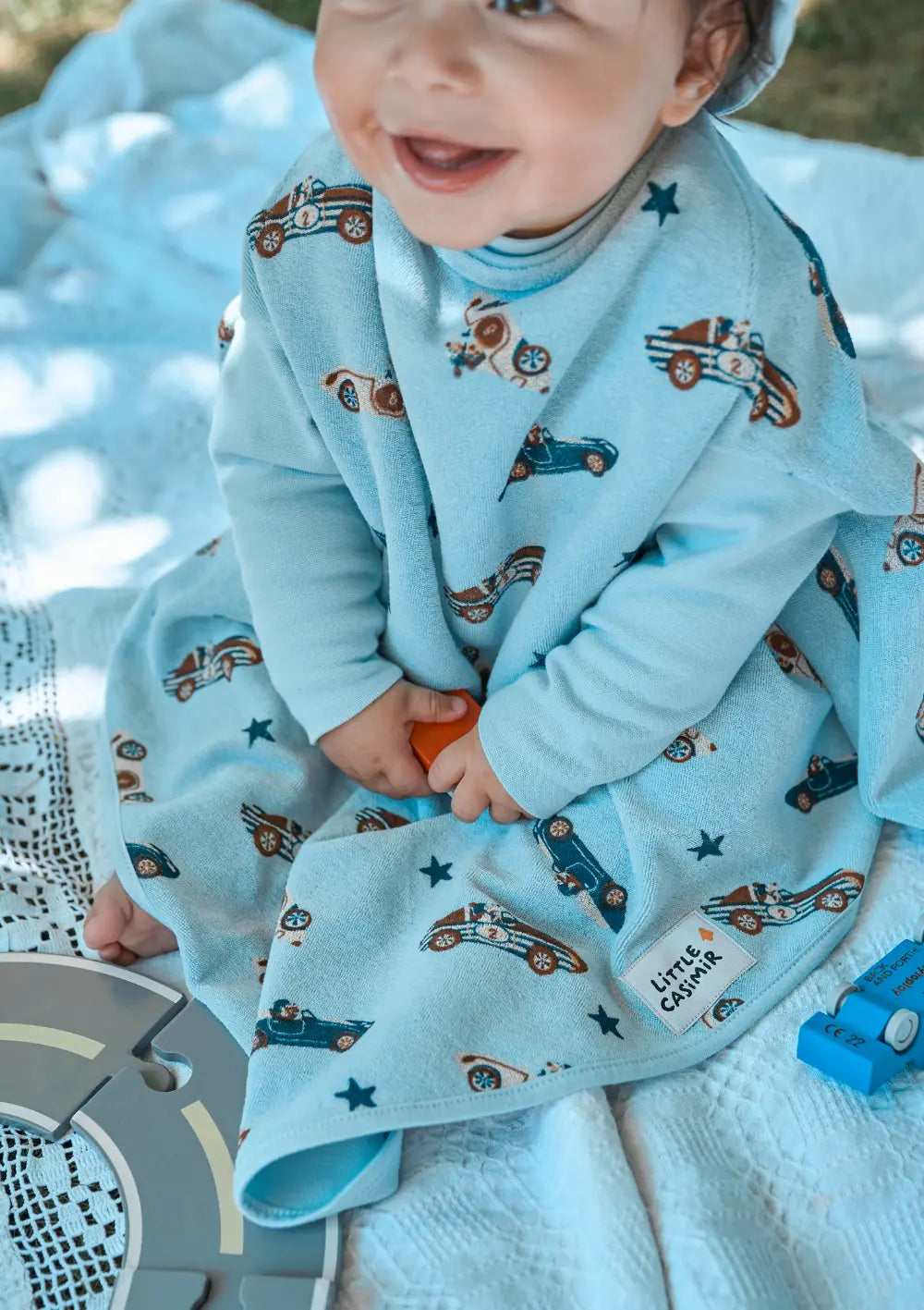 Ein lächelndes Kleinkind sitzt auf einer Decke im Freien und spielt; geschützt wird die Kleidung durch das großzügig geschnittene, hellblaue Little Casimir Langarm-Lätzchen Boo Sky Racers mit Retro-Rennauto-Motiven.
