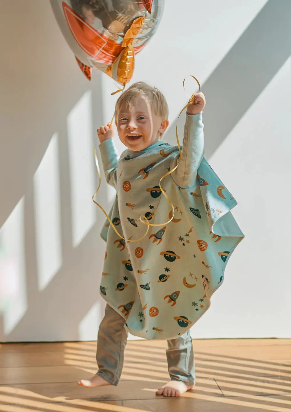 Ein fröhlich lachendes Kind läuft barfuß durch ein lichtdurchflutetes Zimmer und hält die Schnüre eines Raketenballons; es trägt das weite, mintgrüne Little Casimir Langarm-Lätzchen Boo Cosmic 2.0 im Weltraum-Design.