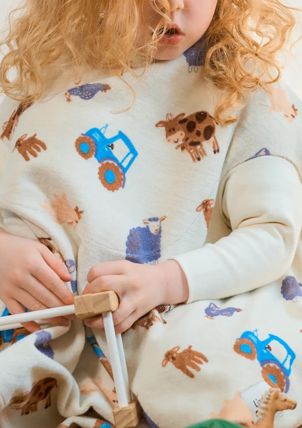 Ein lockiges Kleinkind sitzt beim Spielen auf dem Boden und trägt das großzügig geschnittene Little Casimir Langarm-Lätzchen Boo Farm Friends aus weichem Frottee mit Bauernhof-Motiven.