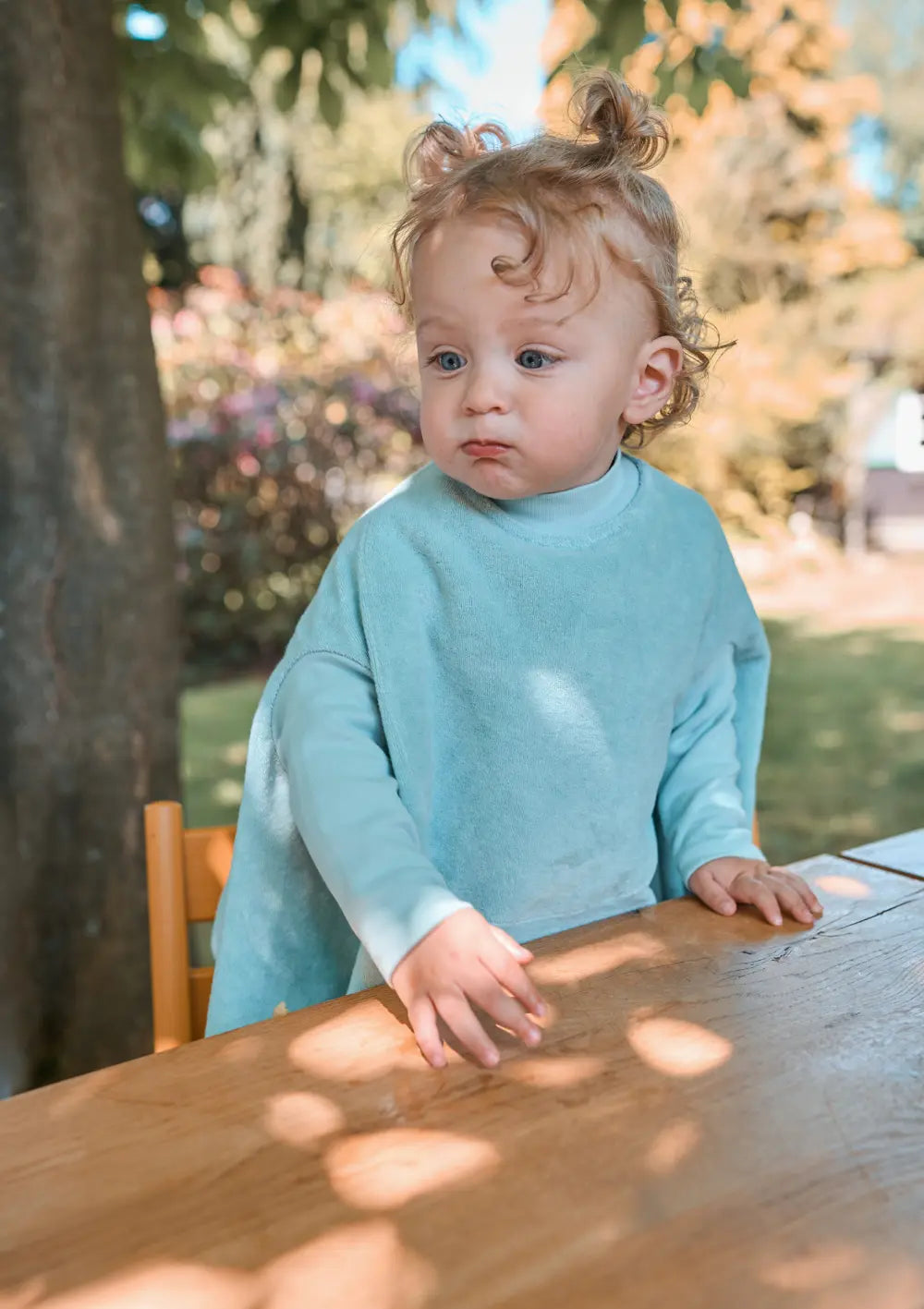 Ein Kleinkind msteht an einem Holztisch im Garten und trägt das mintgrüne Little Casimir Langarm-Lätzchen Boo Matcha Milk aus weichem Frottee