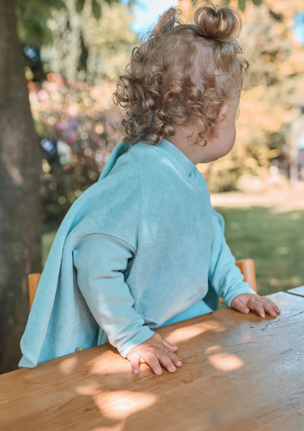 Ein Kleinkind msteht an einem Holztisch im Garten und trägt das mintgrüne Little Casimir Langarm-Lätzchen Boo Matcha Milk aus weichem Frottee