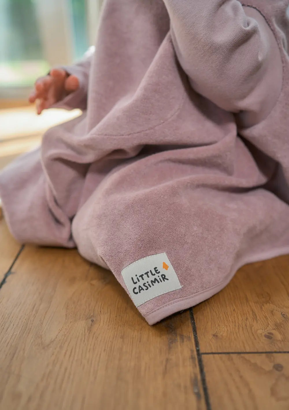 Nahaufnahme auf Holzboden: Der weiche Frottee-Stoff des fliederfarbenen Langarm-Lätzchens Boo Lilac Love mit dem weiß aufgenähten Marken-Label „Little Casimir“.