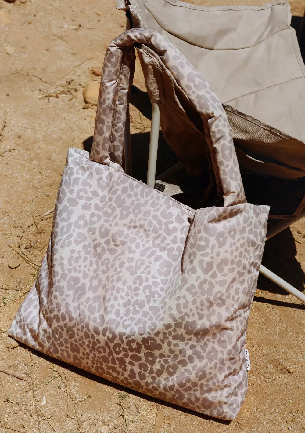 Nahaufnahme der gemusterten Studio Noos Tasche Puffy Mom Bag Beige Leopard; die wattierte, große Schultertasche mit auffälligem Leopardenmuster lehnt im Freien an einem faltbaren Campingstuhl.