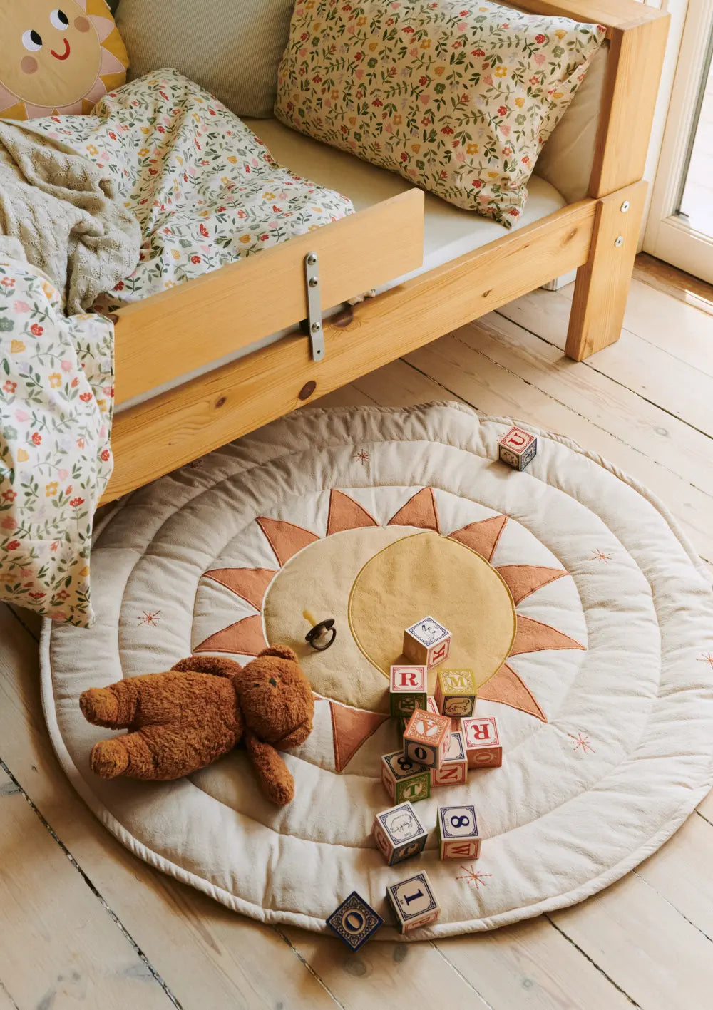Die runde, cremefarbene Dear April Spieldecke Sirius Soft White mit einem großen Sonnen- und Mond-Motiv liegt neben einem Kinderbett auf dem Holzfußboden, dekoriert mit Bausteinen und einem Teddybär.