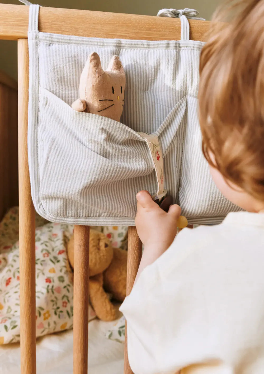 Ein Kleinkind greift nach einem Schnuller an der grau-weiß feingestreiften Dear April Betttasche Polly Dew Stripe, die mit praktischen Aufbewahrungsfächern am Holzgitter des Kinderbettes festgebunden ist.