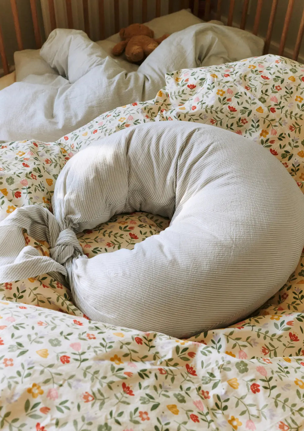 Das ergonomisch geschwungene, grau-weiß gestreifte Dear April Stillkissen Nami Dew Stripe liegt zusammengeknotet auf einer weichen, floral gemusterten Bettwäsche im Babybett.