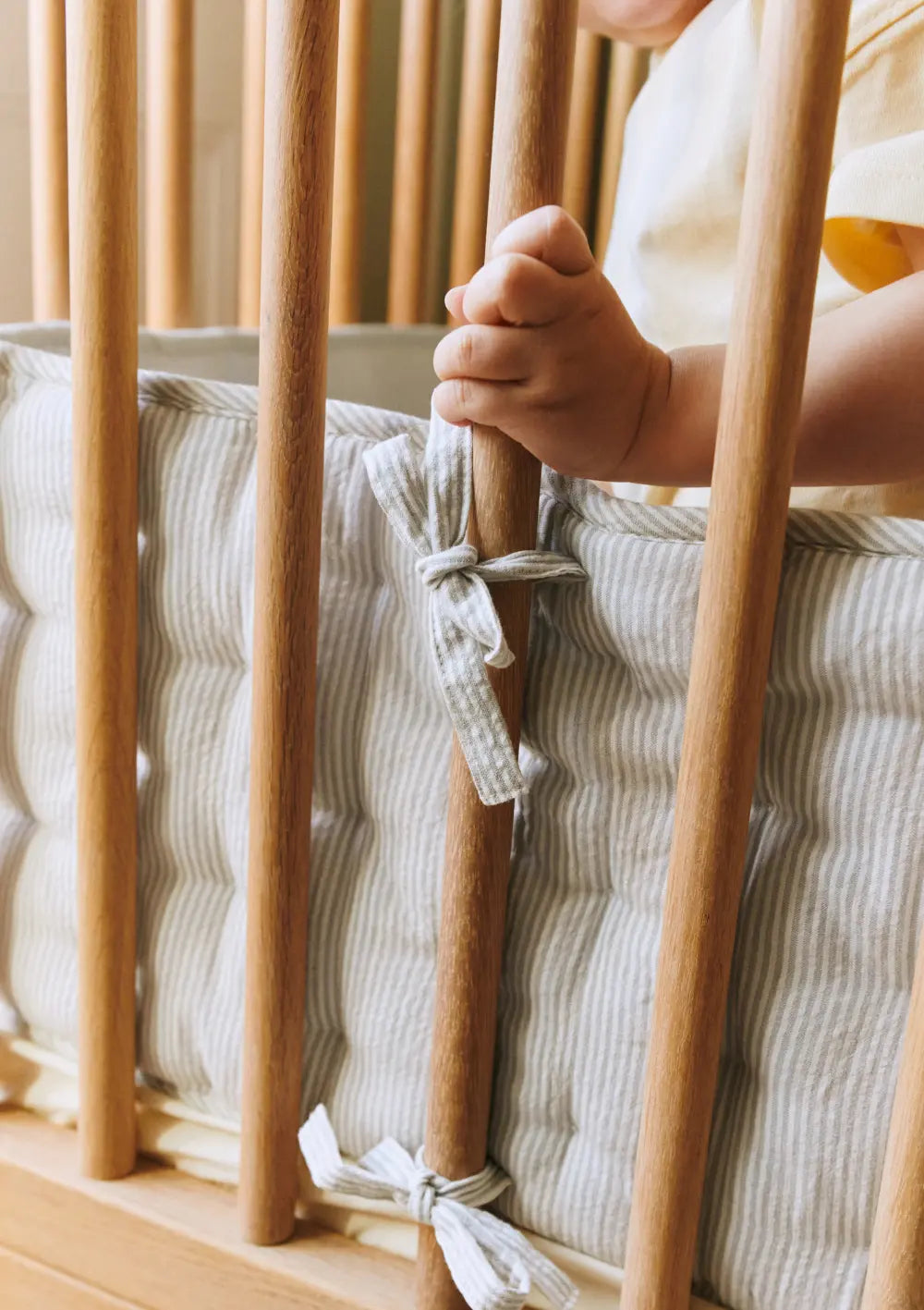 Nahaufnahme eines Babygitterbettes aus Holz; die Hand eines Babys greift nach den Stäben über dem grau-weiß gestreiften, weich gepolsterten Dear April Bettnestchen Nori Dew Stripe.