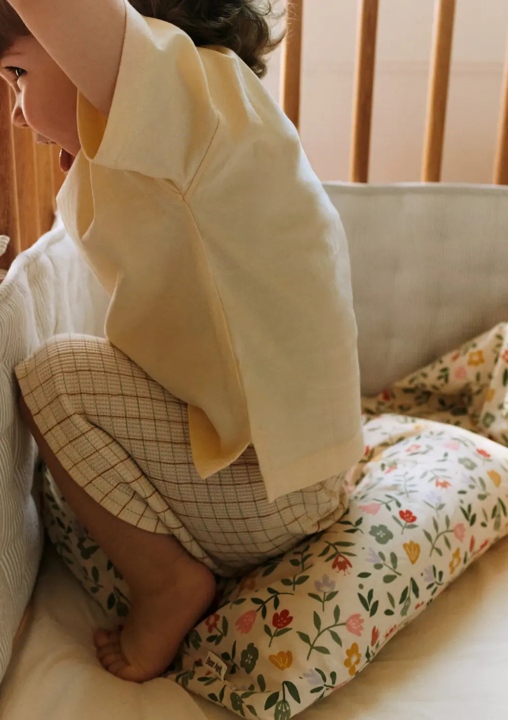 Ein Kleinkind kniet im Gitterbett an der weichen Umrandung durch das grau-weiß gestreifte Dear April Bettnestchen Nori Dew Stripe; im Bett liegt eine bunte Blumen-Bettwäsche.