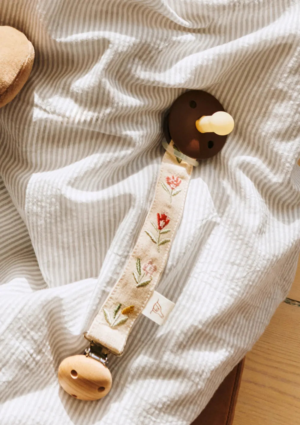 Eine elegante Dear April Schnullerkette Bibi Flower Market aus besticktem Stoff mit Holzclip liegt befestigt an einem braunen Beruhigungssauger auf feingestreiftem Stoff.