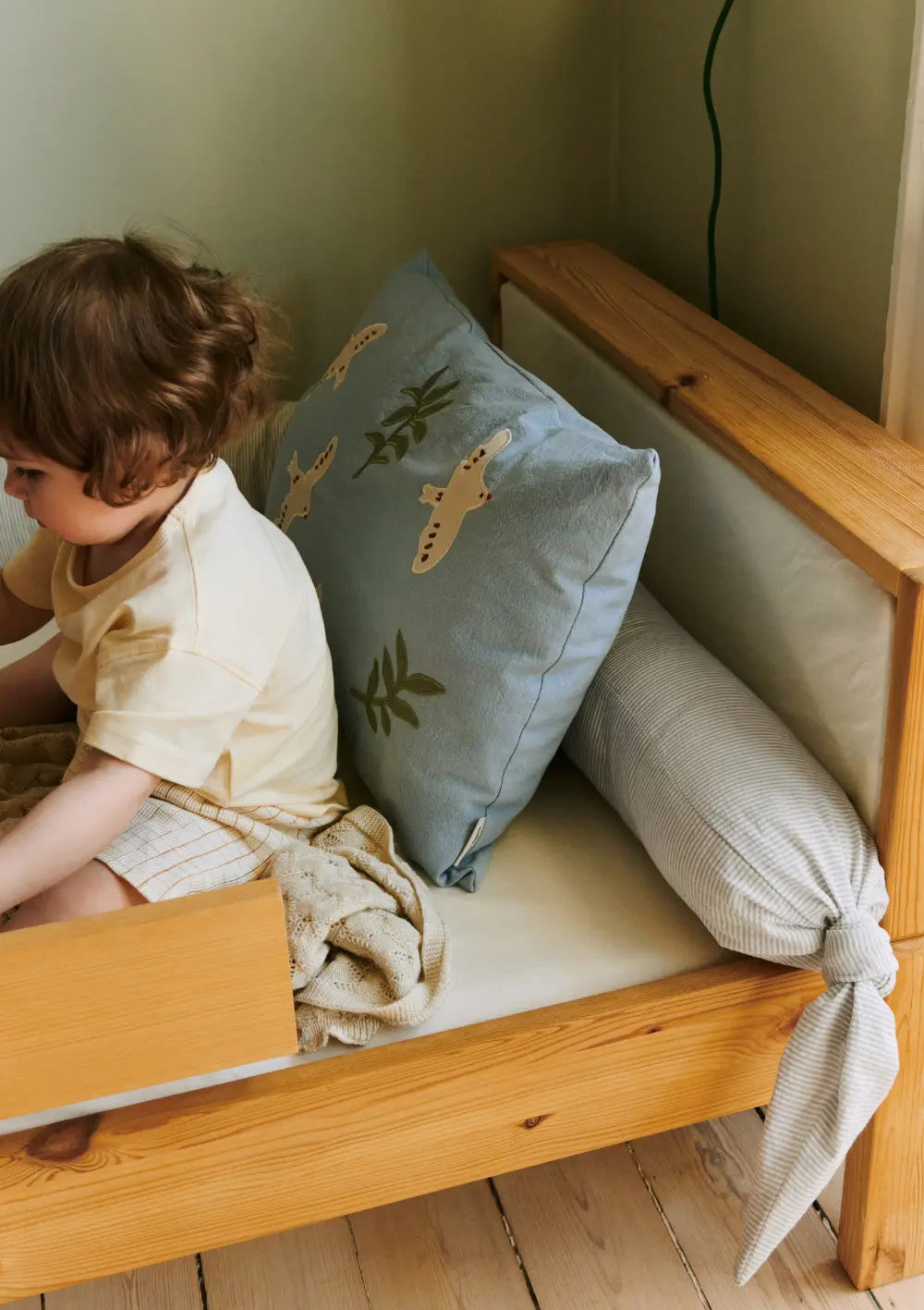 Ein Kleinkind sitzt in einem hölzernen Kinderbett, in dem die grau-weiß gestreifte Dear April Bettschlange Skye Dew Stripe mit dekorativ verknotetem Ende als schützende Nestchen-Polsterung an der Kopfseite liegt.