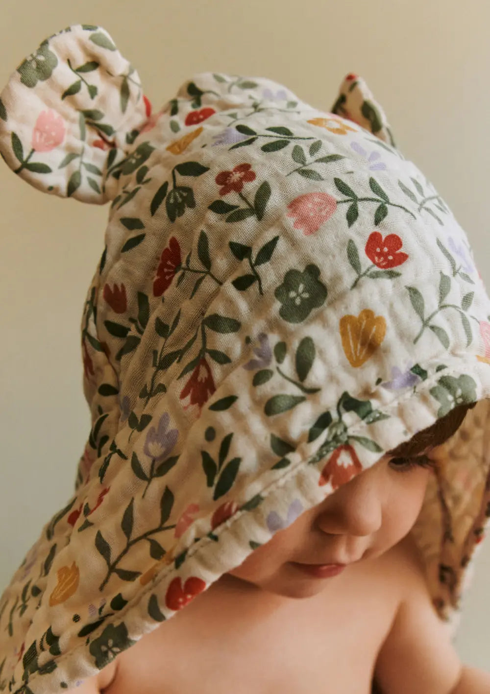 Nahaufnahme eines Babys mit dem Dear April Musselin Kapuzenhandtuch Balou Flower Market, das mit niedlichen angenähten Bärenohren an der floral bedruckten Kapuze gestaltet ist.