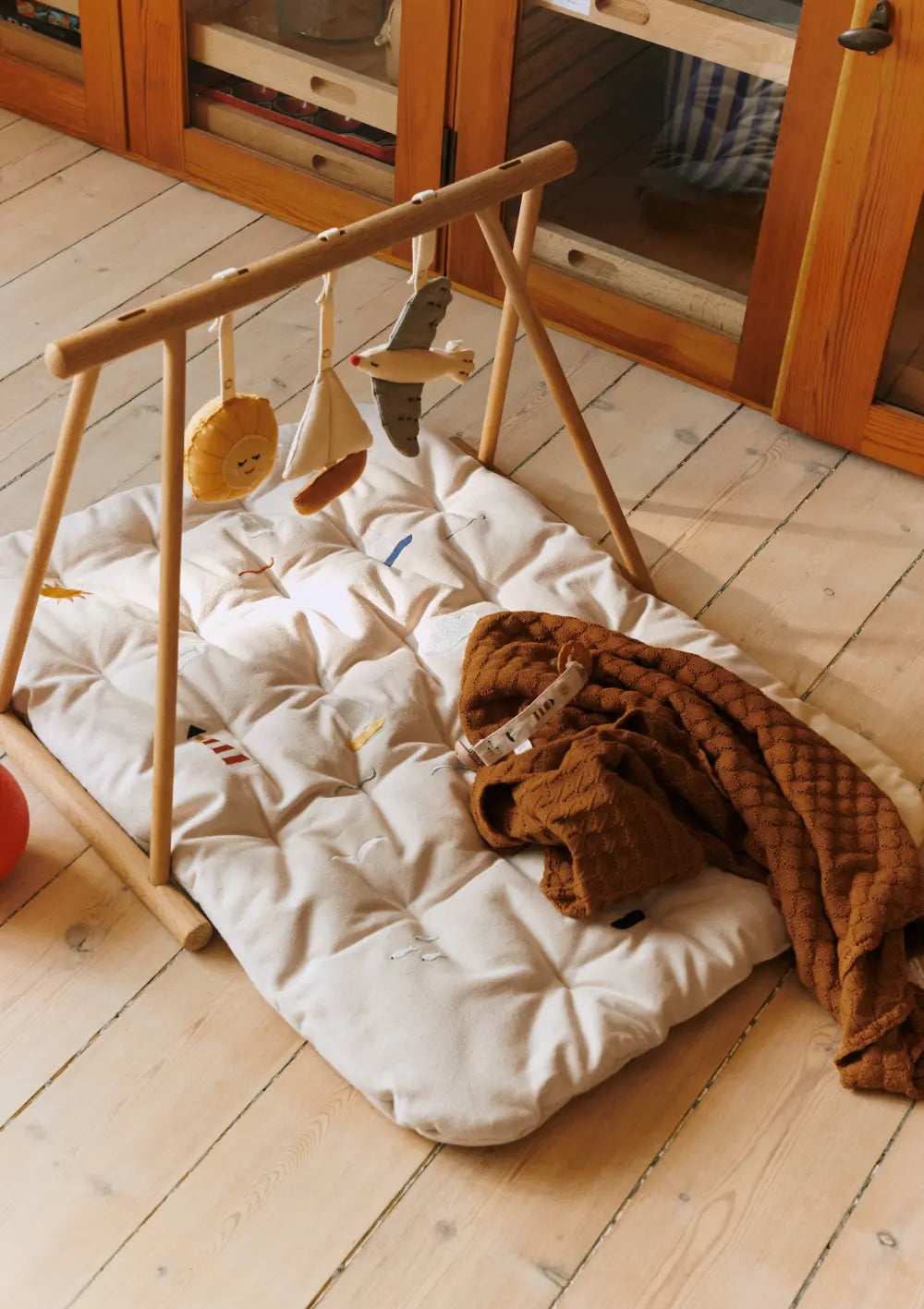 Ein hölzerner Spielbogen mit maritimen Anhängern steht über der beigen, gesteppten Dear April Spieldecke Morris Sea Life, auf der eine braune Strickdecke abgelegt ist.