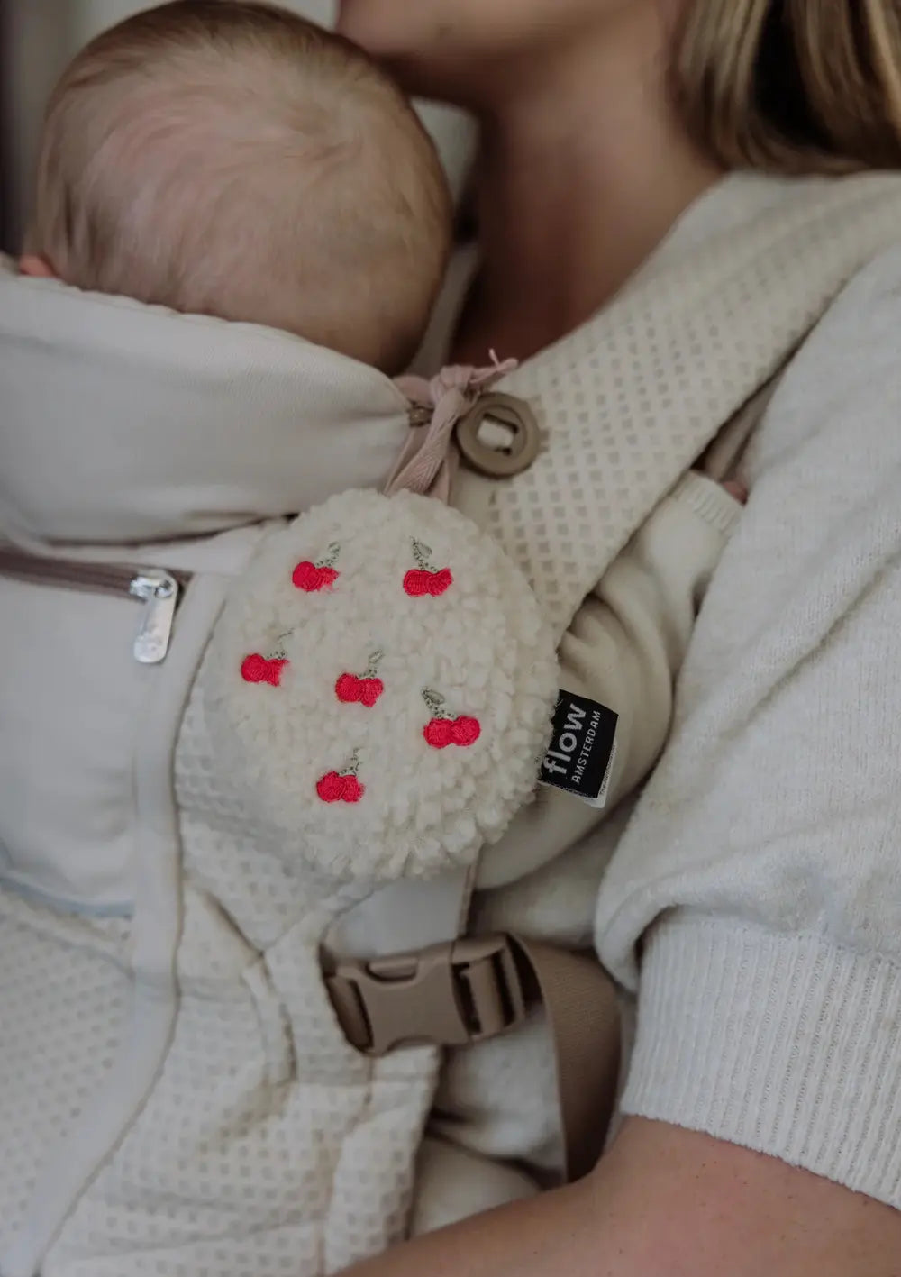Nahaufnahme der runden Flow Amsterdam Einschlafhilfe für unterwegs Lucy Comforter on the go Cherry mit rotem Kirschmuster, die mithilfe einer Stoffschlaufe an einer hellen Babytrage vor der Brust der Mutter befestigt ist.