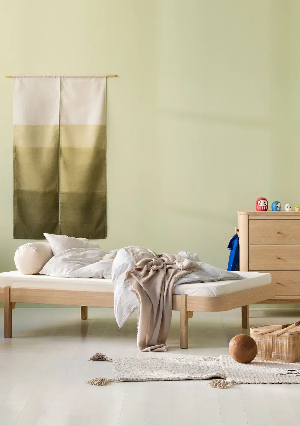 Das elegante Oliver Furniture Lounger Bett Wood Eiche 200 x 120 cm fügt sich mit seiner breiten Liegefläche und hellen Kissen harmonisch vor einer lindgrünen Wand neben der passenden Holzkommode ein.