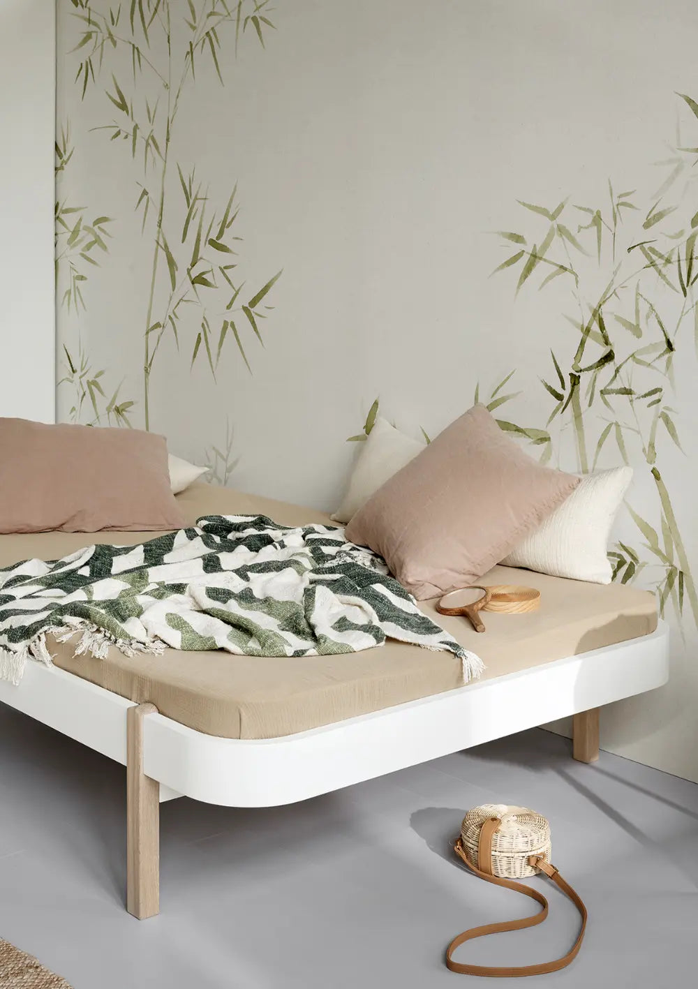 Das flache Oliver Furniture Wood Lounger Bett weiß eiche präsentiert sich mit einem weiß lackierten Bettrahmen und abgerundeten Ecken sowie Bettbeinen aus natürlichem Eichenholz vor einer Wand mit dezentem Bambus-Dekor.