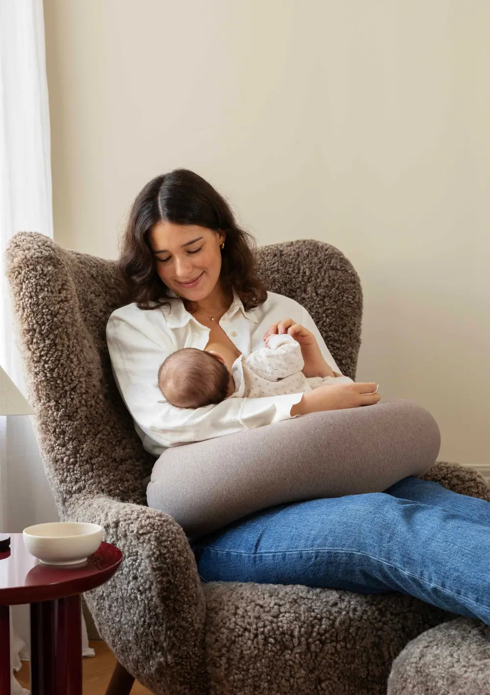 Eine lächelnde Mutter sitzt in einem gemütlichen, braunen Teddy-Sessel und nutzt das u-förmige bbhugme Stillkissen Hazel Brown auf ihrem Schoß zur komfortablen Entlastung beim Stillen ihres Babys.