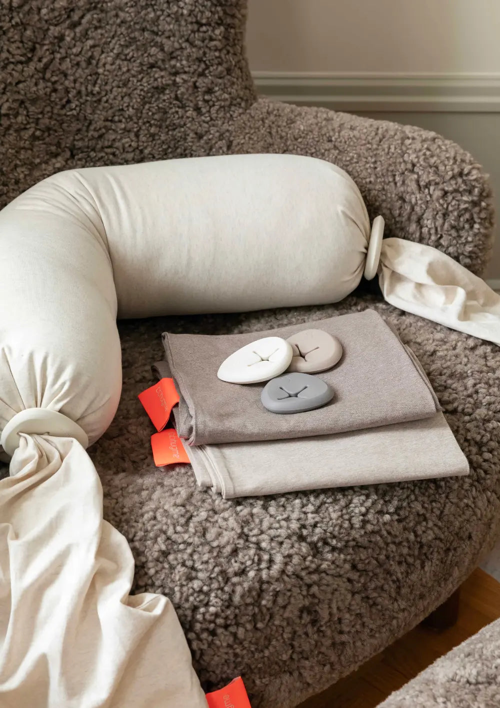 Das cremefarbene bbhugme Stillkissen Soft White liegt auf einem braunen Plüschsessel neben gefalteten Ersatzbezügen und farblich passenden Pebble-Silikonringen zum Abbinden der Kissenenden.