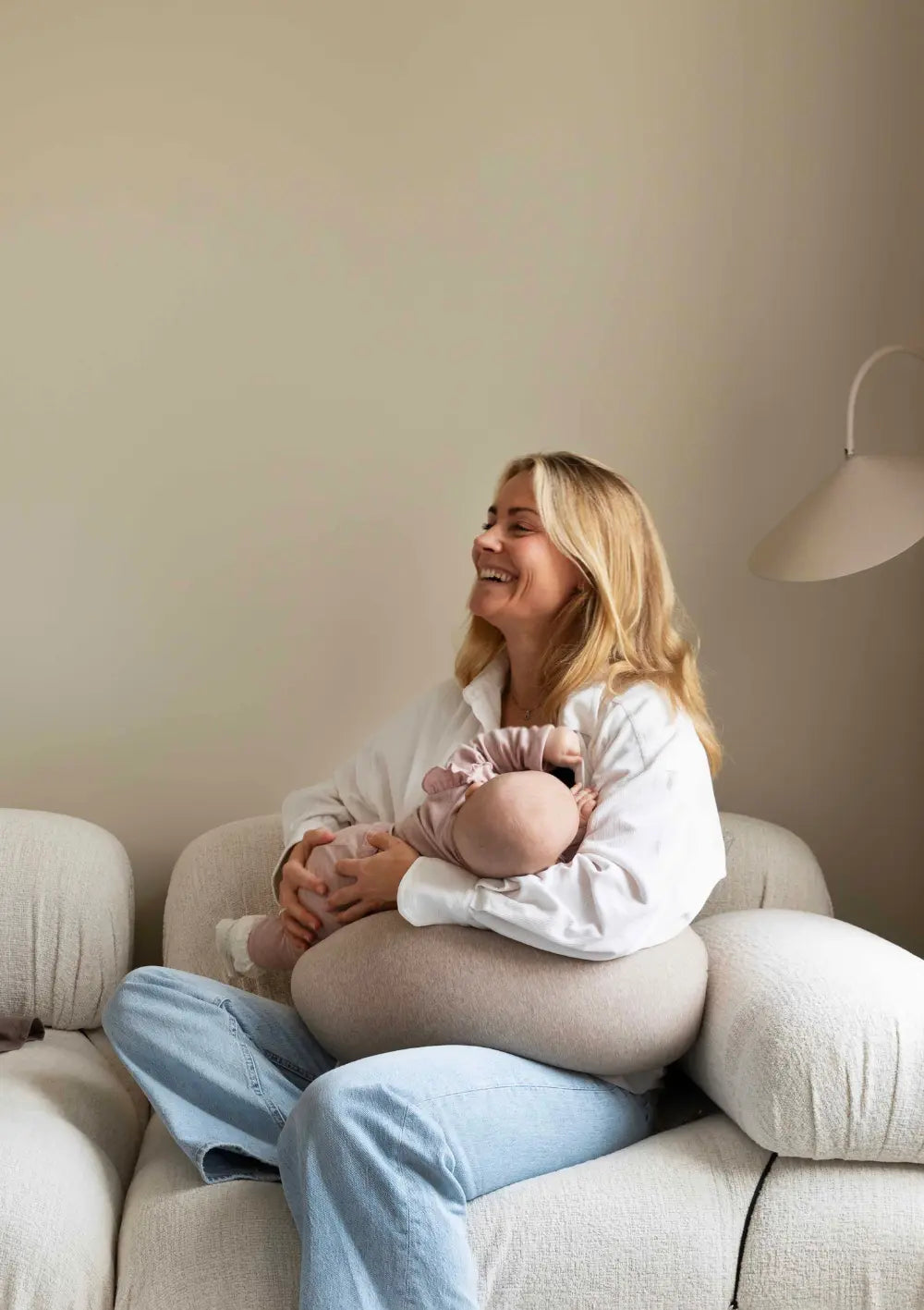 Eine glücklich lachende Mutter sitzt auf einem hellen Bouclé-Sofa und hält ihr Baby im Arm, das gemütlich auf dem u-förmig angepassten, sandfarbenen bbhugme Stillkissen Seashell Beige auf ihrem Schoß gebettet ist.