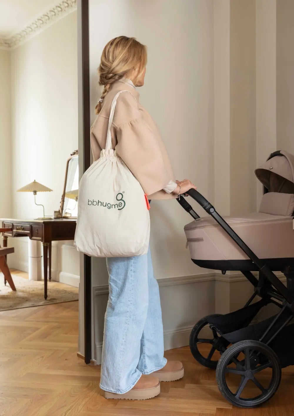 Eine Frau steht mit einem Kinderwagen im Flur und trägt die weiße Stoff-Tragetasche mit dem grünen Marken-Logo des bbhugme Stillkissen Seashell Beige über der Schulter, um die einfache Transportierbarkeit zu zeigen.