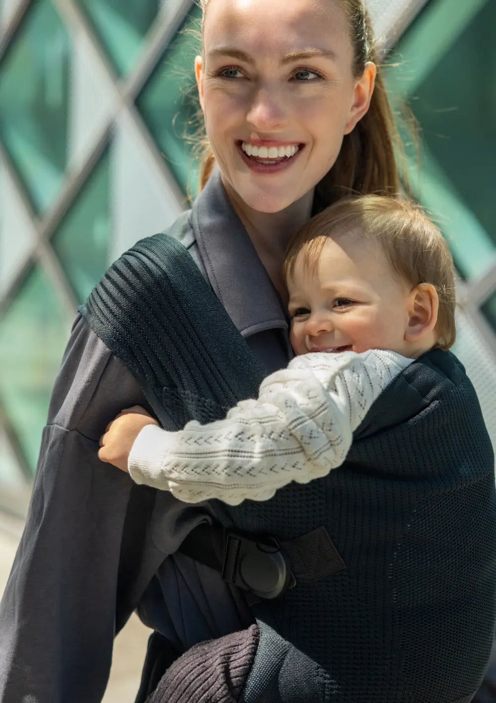 Eine glückliche Mutter trägt ihr lächelndes Kleinkind in der komfortablen Flybaby Kleinkindtrage Black unterwegs vor dem Bauch; die Detailaufnahme zeigt das anpassungsfähige Tragesystem mit Sicherheitsverschluss.
