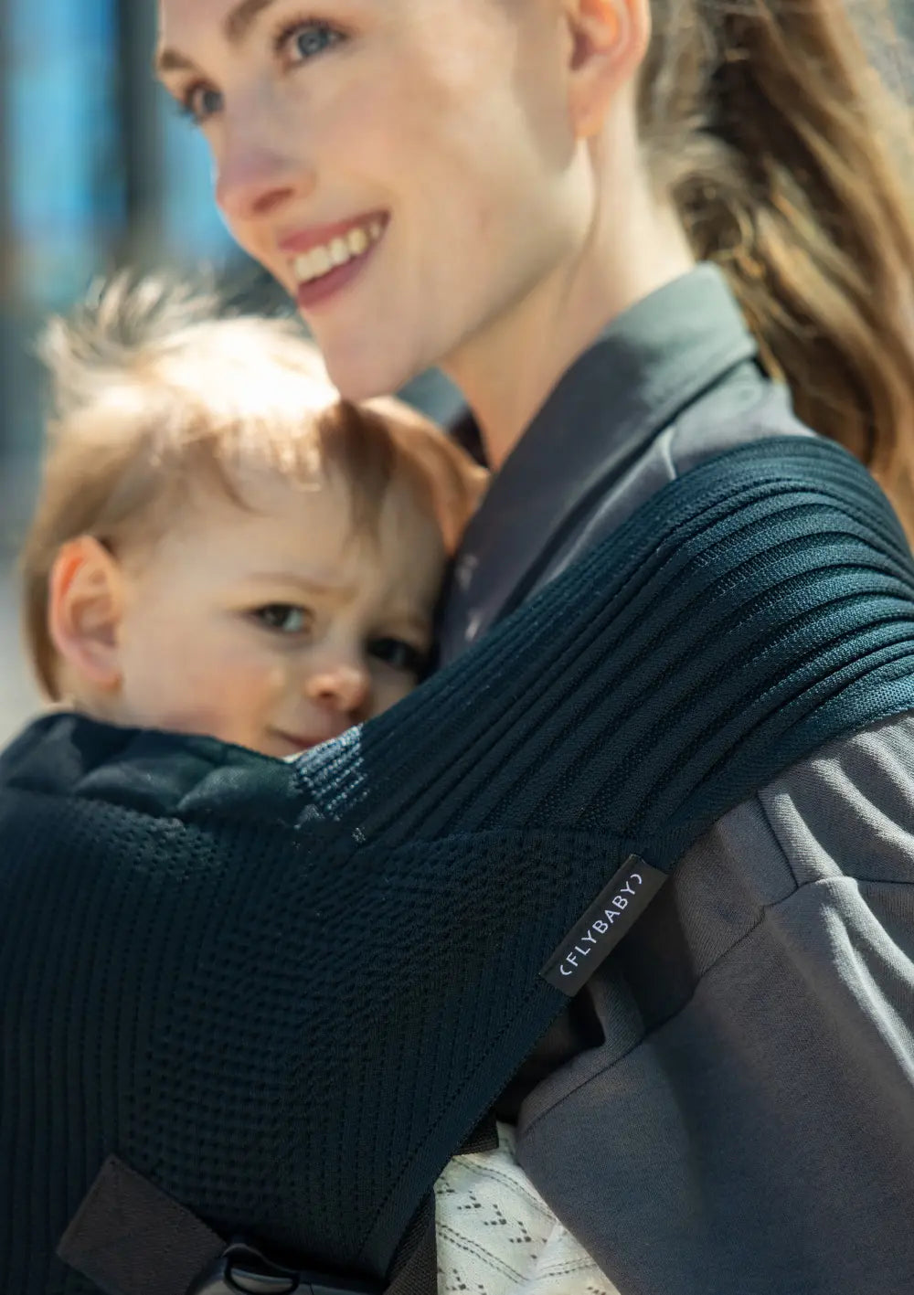 Nahaufnahme einer lächelnden Mutter, die ihr Kleinkind in der ergonomischen Flybaby Kleinkindtrage Black vor der Brust trägt; der Fokus liegt auf der weich gestrickten, schwarzen Oberflächenstruktur des breiten Schultergurts.