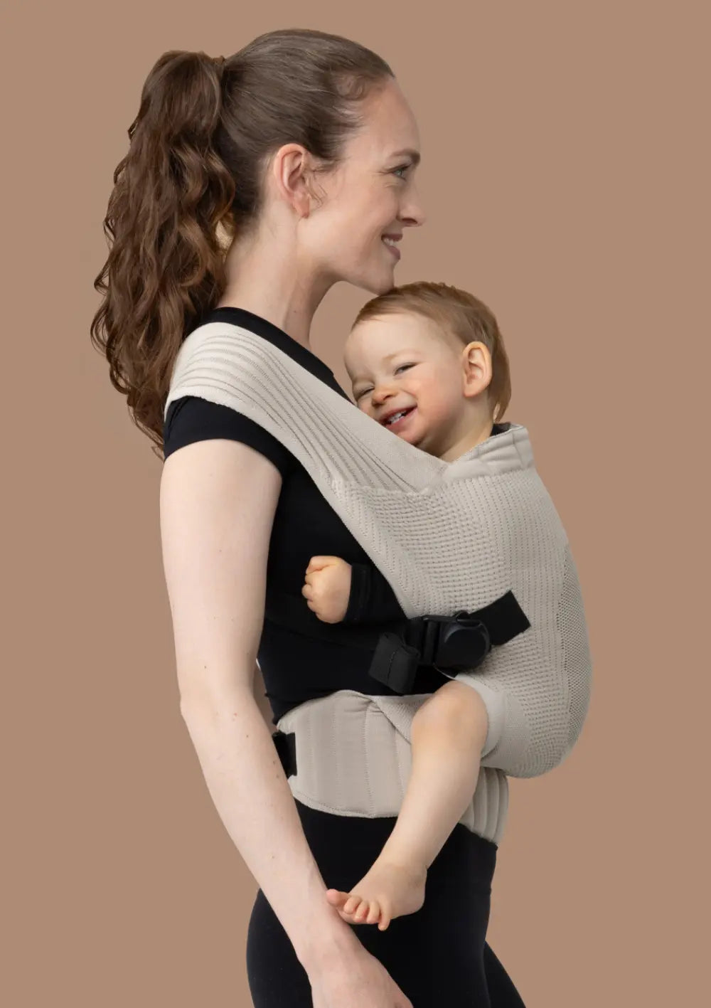 Frau trägt ihr Kleinkind in der Flybaby Kleinkindtrage Creme vor dem Bauch.
