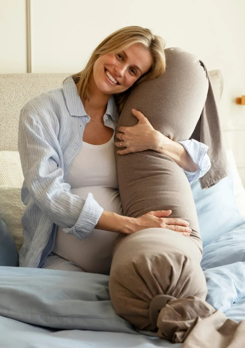 Eine glücklich lächelnde Schwangere sitzt im Bett und hält das flexible bbhugme Schwangerschaftskissen Hazel Brown innig im Arm; im Vordergrund ist das mit einem Pebble-Silikonring dekorativ abgebundene Ende gut sichtbar.