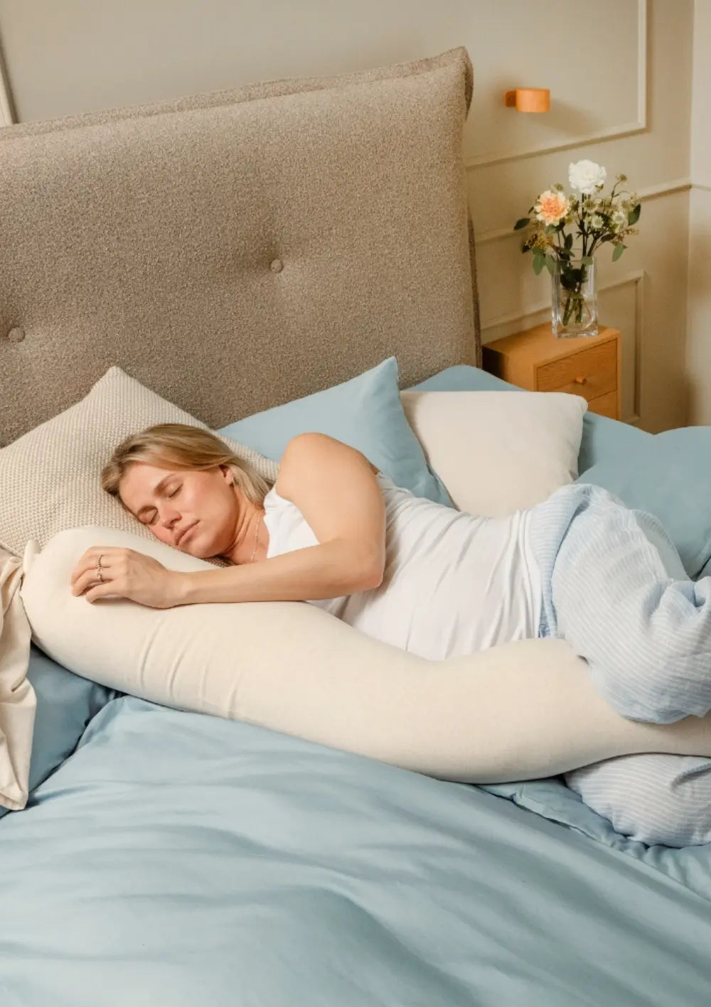Eine schwangere Frau schläft entspannt in Seitenlage im Bett und nutzt das lange, cremefarbene bbhugme Schwangerschaftskissen Soft White zur optimalen Entlastung von Bauch und Knien.