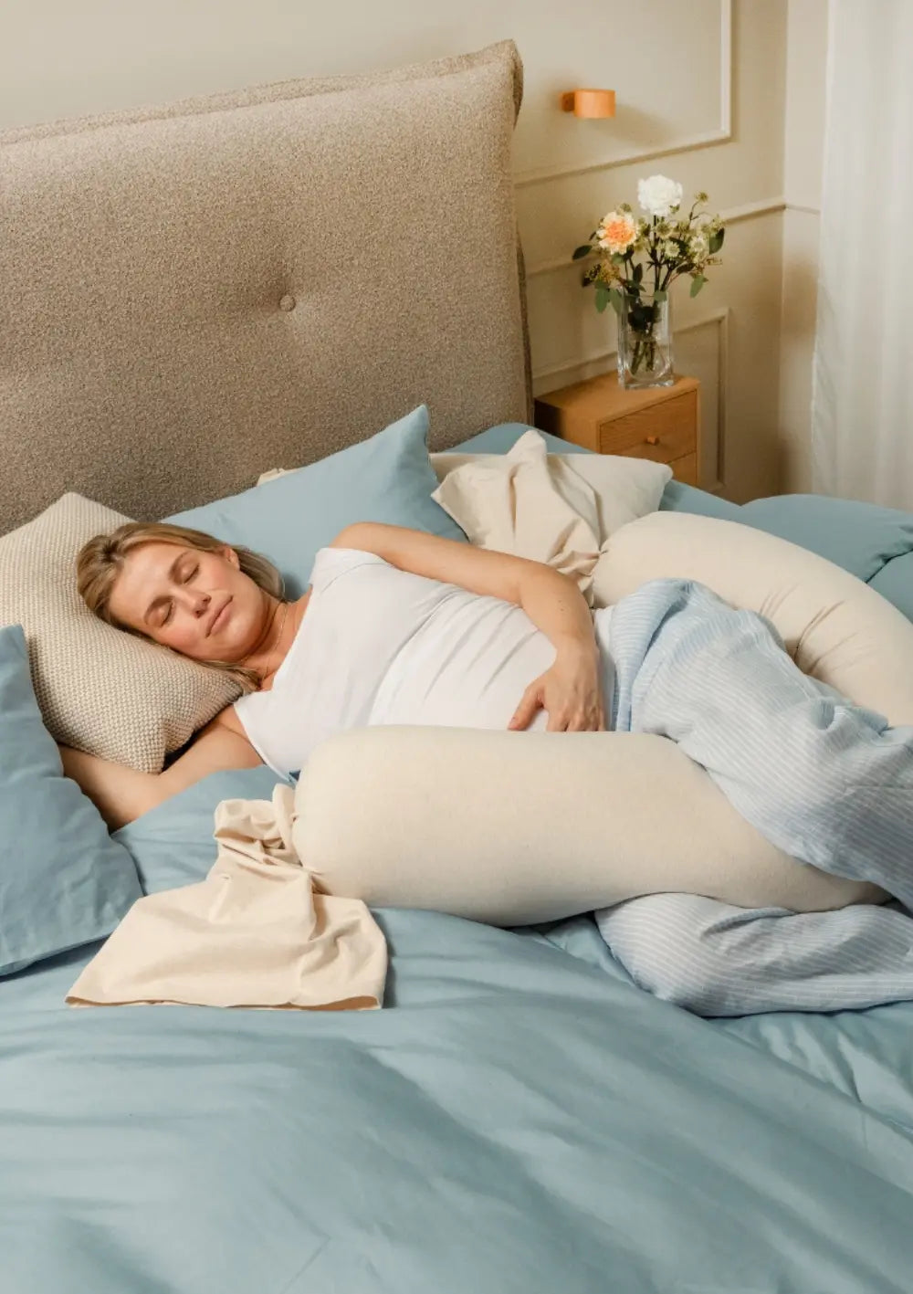 Eine Schwangere ruht in Seitenlage im Bett und stabilisiert ihren Körper mit dem flexibel geschwungenen, weichen bbhugme Schwangerschaftskissen Soft White.