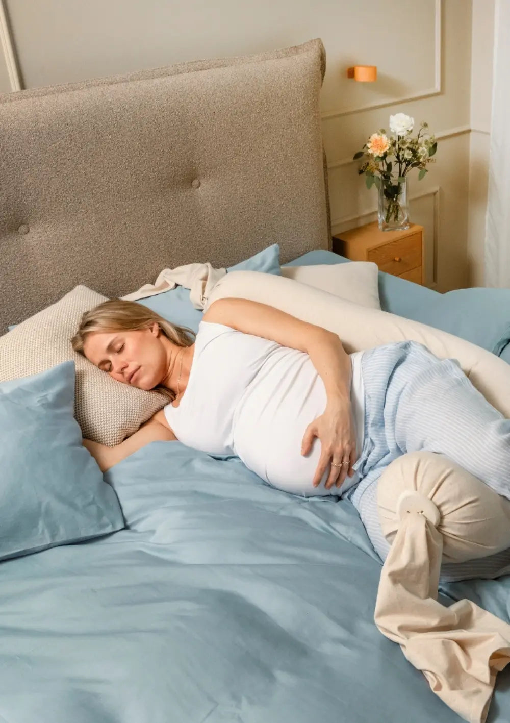 Eine schwangere Frau schläft ruhig auf der Seite im Bett, während das lange bbhugme Schwangerschaftskissen Soft White hinter ihrem Rücken platziert ist, um eine stabile und bequeme Schlafposition zu unterstützen.