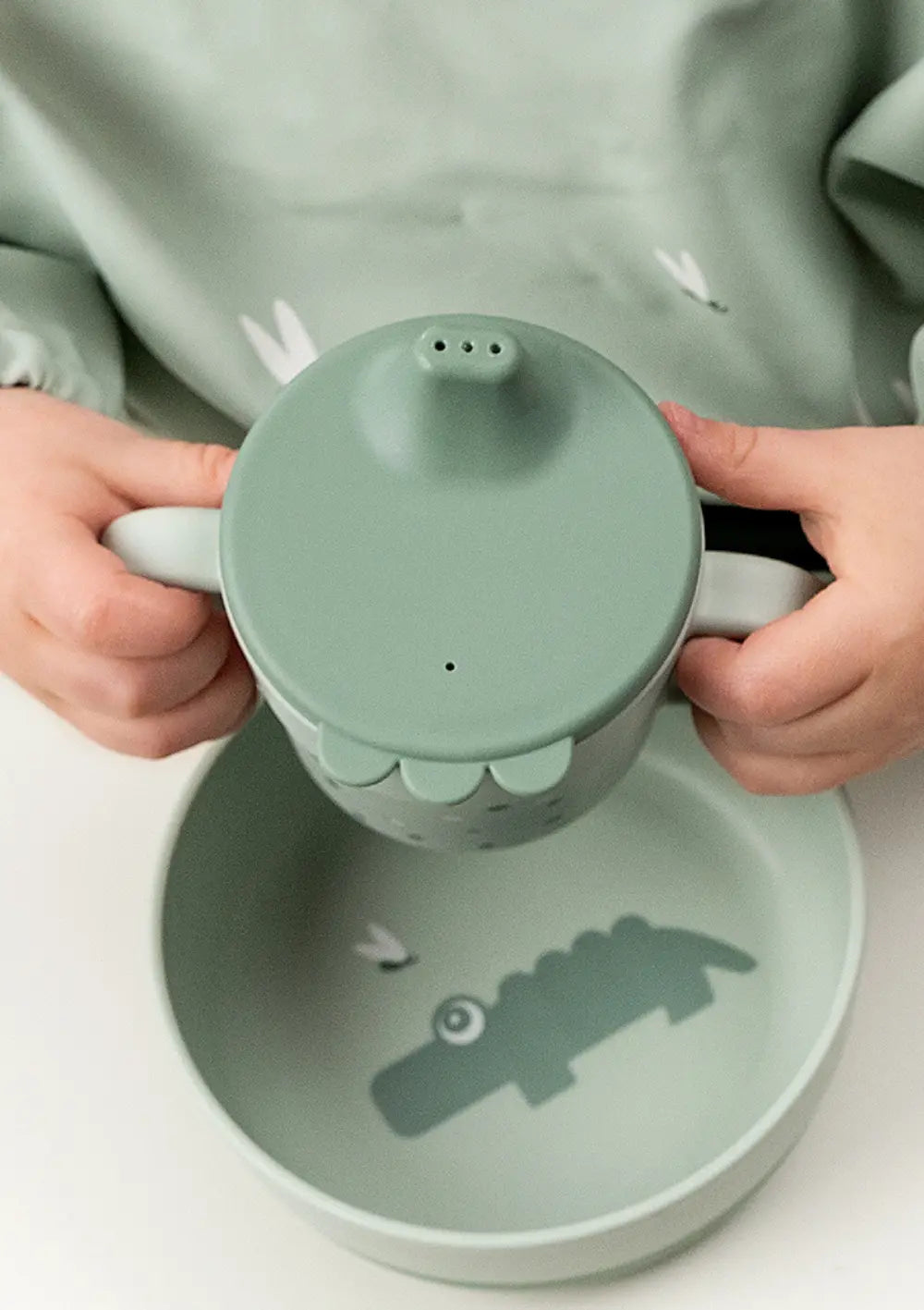 Done by Deer Trinklernbecher 'Foodie' Happy dots grün wird von zweil kleinen Kinderhänden an den Henkeln festgehalten