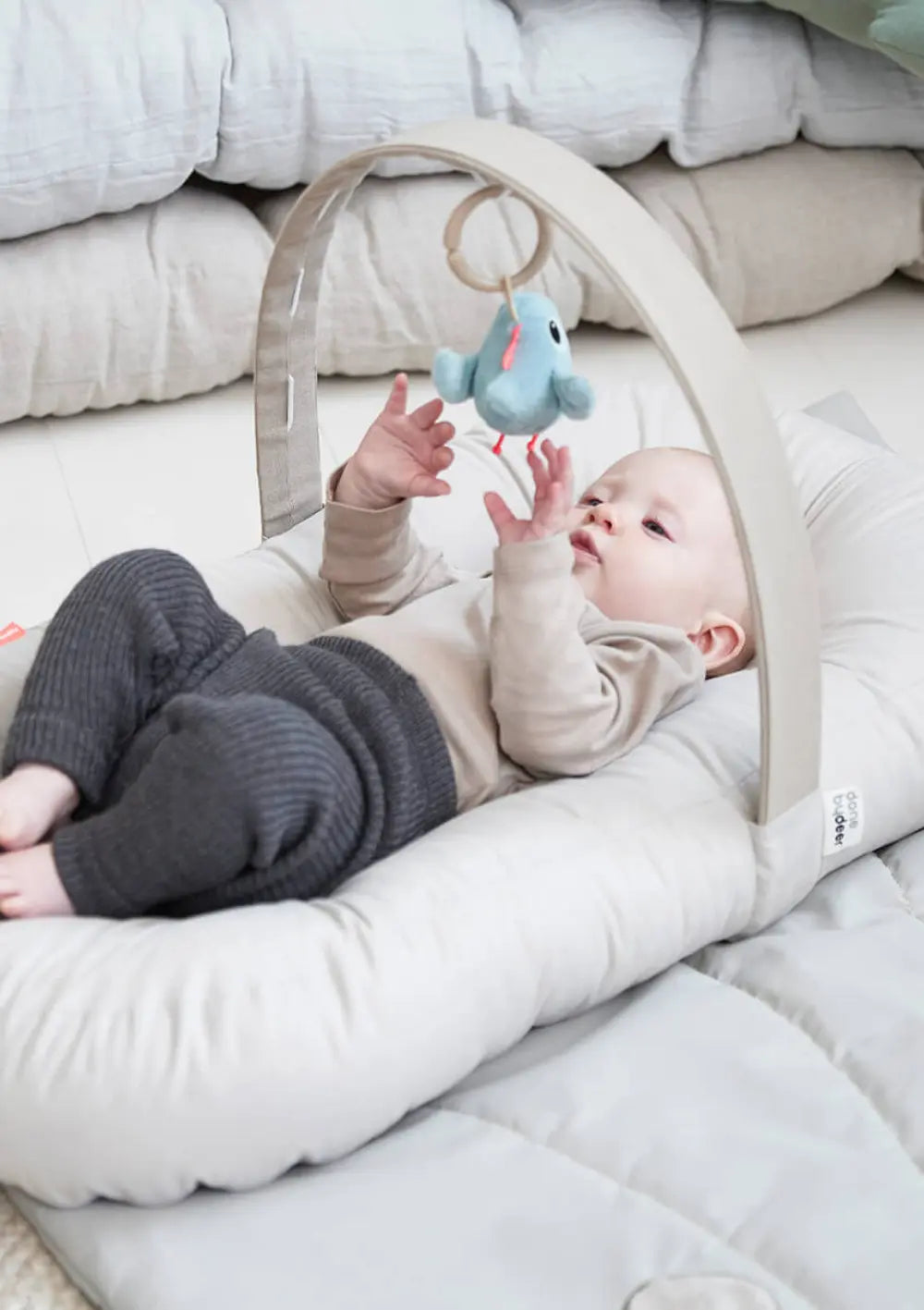 in Baby liegt geborgen auf dem sandfarbenen Done by Deer Babykissen mit Aktivitätsbogen Cozy Lounger Raffi Sand und streckt die Hände nach einem kleinen, blauen Plüschvogel aus, der am Spielbogen hängt.