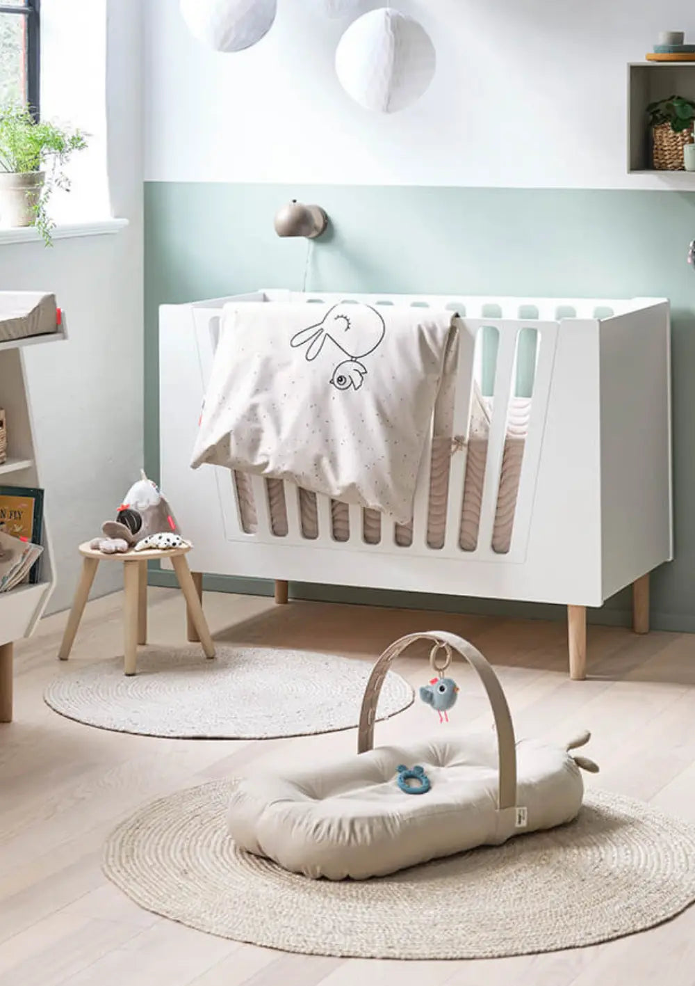 Das sandfarbene Done by Deer Babykissen mit Aktivitätsbogen Cozy Lounger Raffi Sand liegt aufgebaut auf einem runden Juteteppich im Kinderzimmer vor einem weißen Gitterbett.
