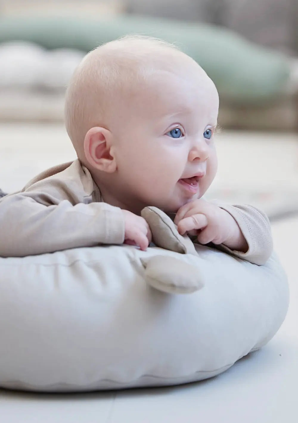 Ein lächelndes Baby trainiert die Bauchlage und stützt sich mit den Unterarmen auf der weich gepolsterten Umrandung des Done by Deer Babykissen mit Aktivitätsbogen Cozy Lounger Raffi Sand ab.