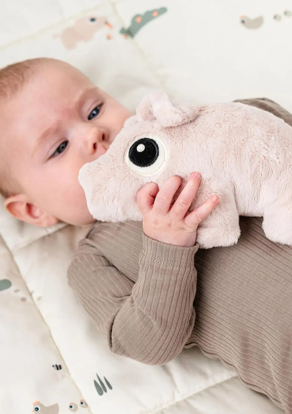 Ein liegendes Baby hält das weiche, hellrosa Done by Deer Kuscheltier Pigee Rosa dicht an seinen Körper geschmiegt; das plüschige Schweinchen ist mit einem großen, gestickten Kullerauge gestaltet.