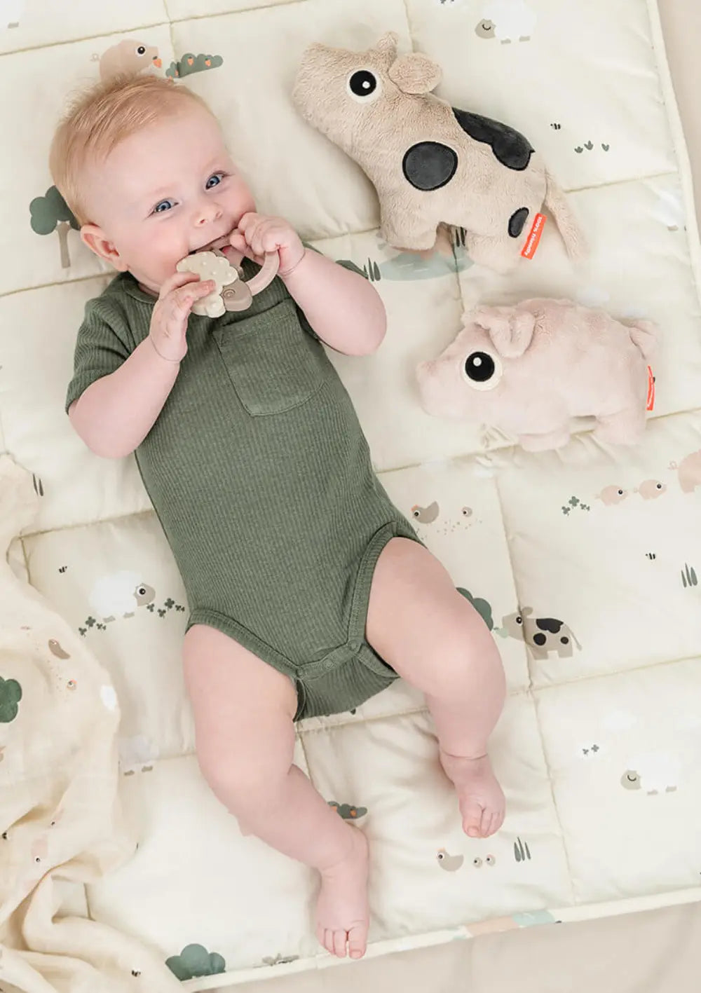 Ein lächelndes Baby liegt auf dem Rücken auf einer gemusterten Spieldecke, nutzt einen Beißring und ist umgeben von zwei Done by Deer Plüschtieren, darunter das kleine, zartrosa Done by Deer Kuscheltier Pigee Rosa.