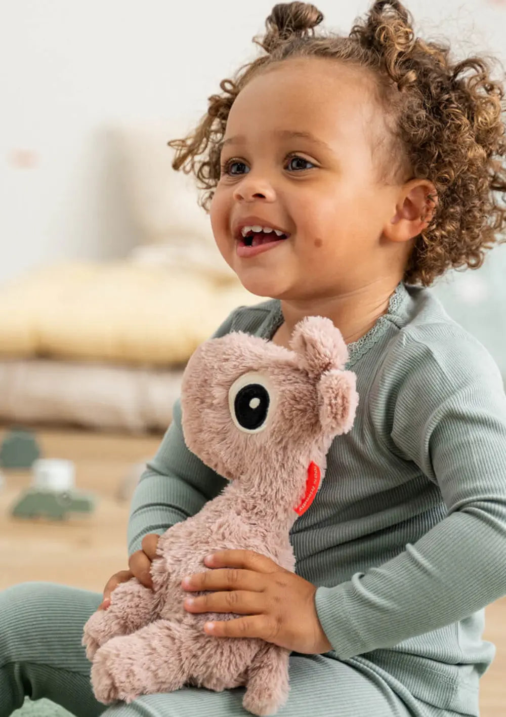 Ein fröhlich lächelndes kleines Mädchen mit Locken sitzt auf dem Zimmerboden und hält die plüschige, altrosa Giraffe Done by Deer Kuschelfreund Raffi Rosa liebevoll im Arm.