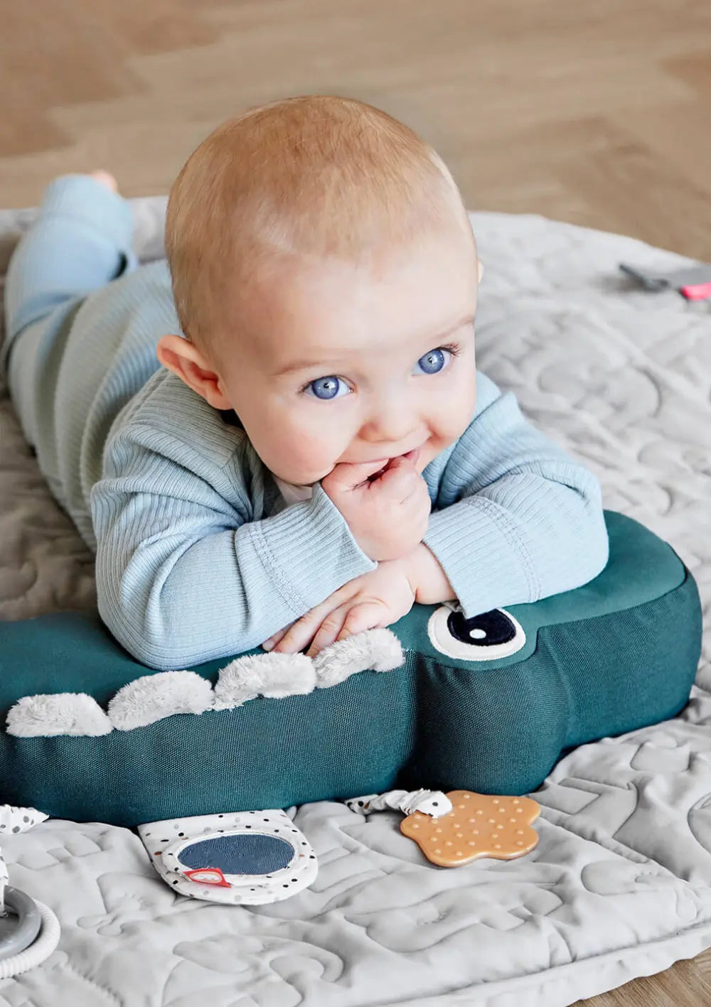 Ein aufgewecktes Baby trainiert die Bauchlage auf einer grauen Krabbeldecke und stützt sich dabei bequem auf dem krokodilförmigen Done by Deer Spielkissen Croco Grün ab.