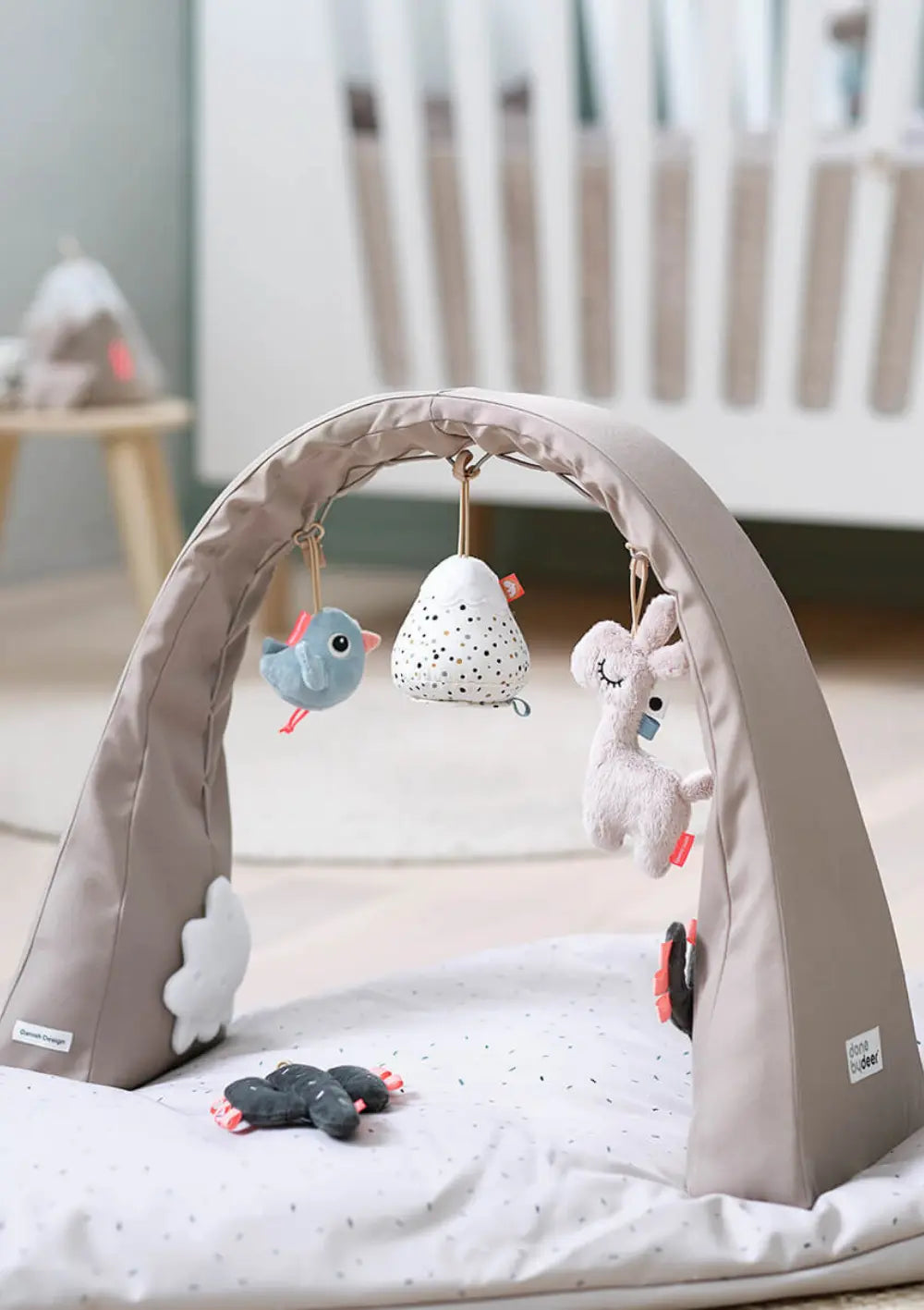 Der freistehende, sandfarbene Done by Deer Spielbogen Activity Gym Lalee Sand steht auf einer gepunkteten Krabbeldecke im Kinderzimmer und ist mit drei hängenden Aktivitätsspielzeugen bestückt.